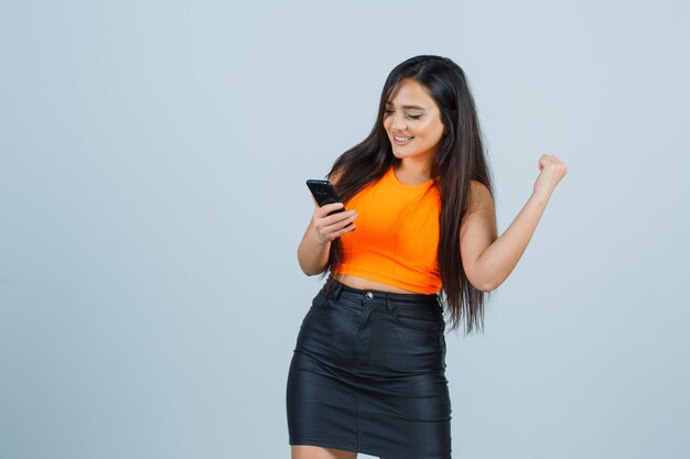 Jovem, levantando o punho enquanto olha para o celular em camiseta, minissaia e parece feliz, vista frontal.