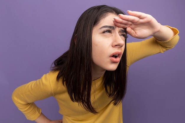 Jovem, linda, caucasiana, colocando a mão na testa, apertando os olhos, olhando para um lado distante, mantendo a outra mão na cintura, isolada na parede roxa