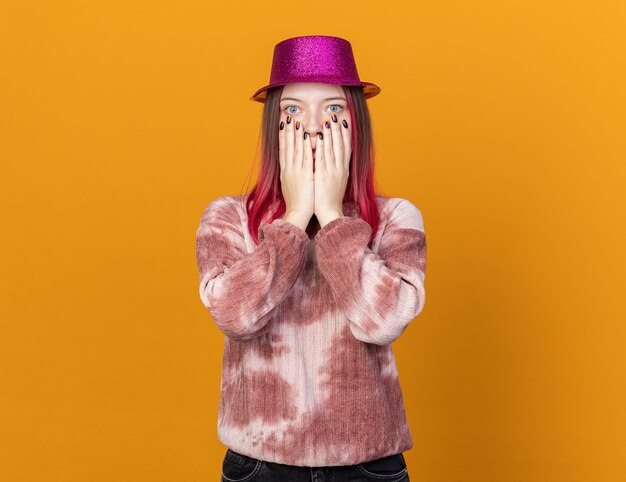 Foto grátis jovem linda com medo de usar chapéu de festa e cobrir o rosto com as mãos isoladas em uma parede laranja