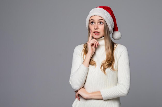 Jovem linda com um chapéu de Papai Noel sobre uma parede branca pensando e olhando para o lado