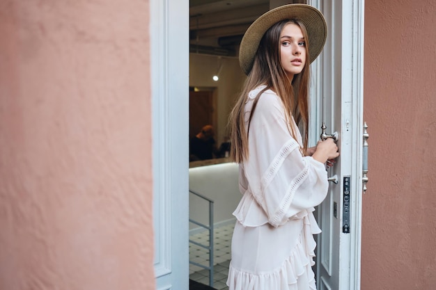 Jovem linda de vestido branco e chapéu olhando pensativamente na câmera enquanto abre a porta branca do café