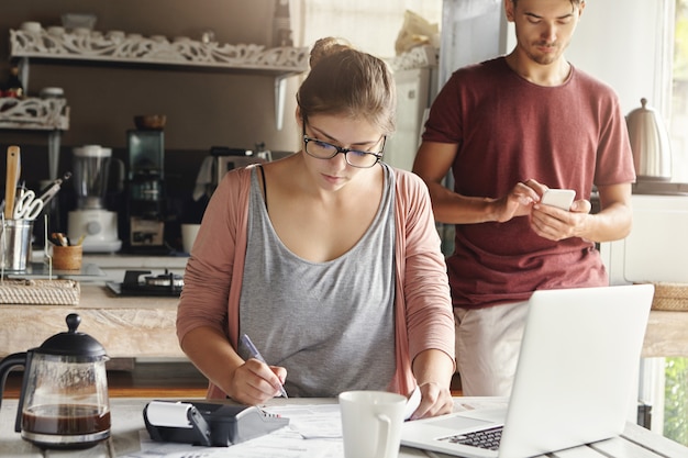 Jovem linda dona de casa usando óculos retangulares, fazendo os cálculos necessários e escrevendo com caneta, enquanto paga contas de serviços públicos, sentado à mesa da cozinha com laptop genérico e calculadora