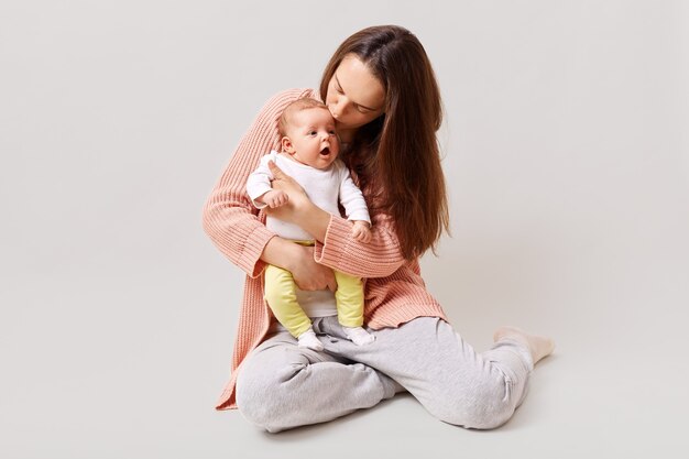 Jovem linda e atraente mãe segurando e beijando um bebê recém-nascido sentado no chão