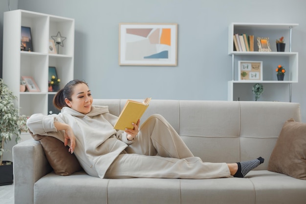 Jovem linda em roupas de casa relaxando em um sofá no interior de casa lendo um livro feliz e positivo sorrindo alegremente