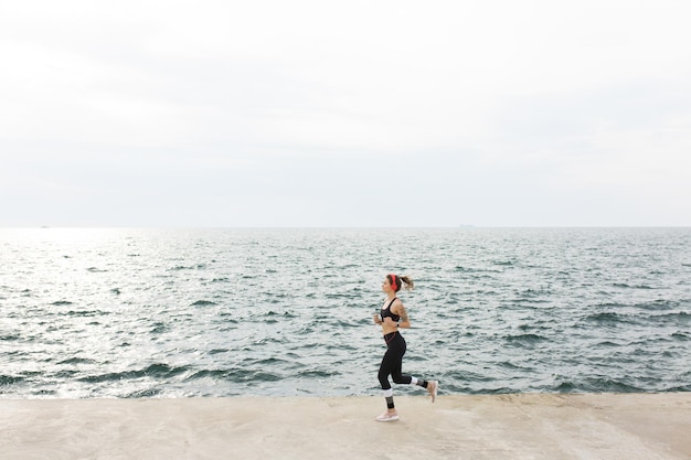 Jovem linda em top esportivo cinza escuro e leggings com fones de ouvido vermelhos correndo sonhadoramente com incrível vista para o mar no fundo