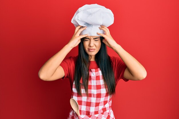 Jovem linda garota hispânica vestindo uniforme de padeiro e chapéu de cozinheiro sofrendo de dor de cabeça desesperada e estressada porque dor e enxaqueca. mãos na cabeça.