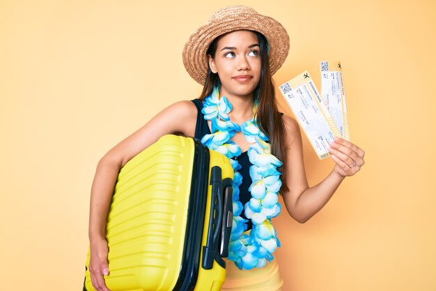 Jovem linda garota latina usando chapéu de verão e lei havaiana segurando mala de cabine e cartão de embarque sorrindo olhando para o lado e olhando para longe pensando.