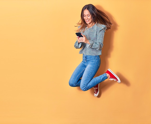 Jovem linda loira pulando com um sorriso no rosto. Usando smartphone sobre fundo amarelo isolado