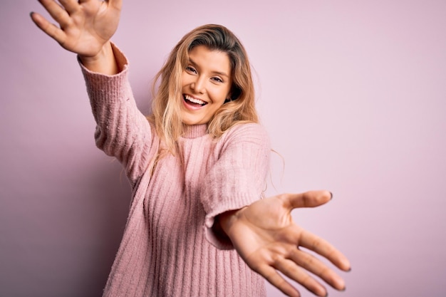 Jovem linda loira vestindo suéter rosa casual sobre fundo isolado, olhando para a câmera, sorrindo de braços abertos para o abraço Expressão alegre, abraçando a felicidade