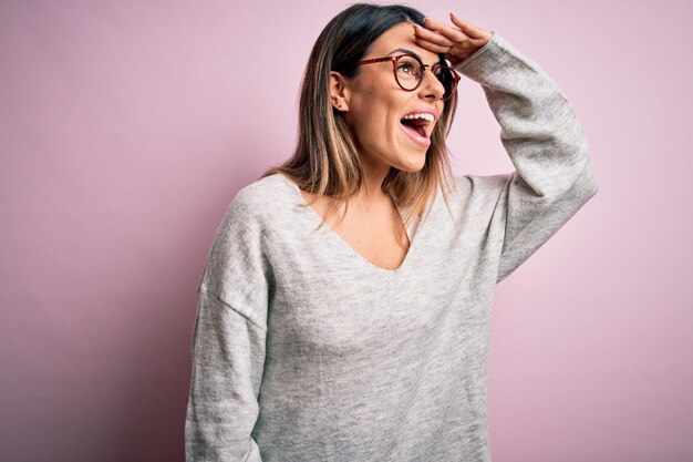 Jovem linda morena vestindo suéter casual e óculos sobre fundo rosa muito feliz e sorrindo olhando para longe com a mão sobre a cabeça Conceito de pesquisa