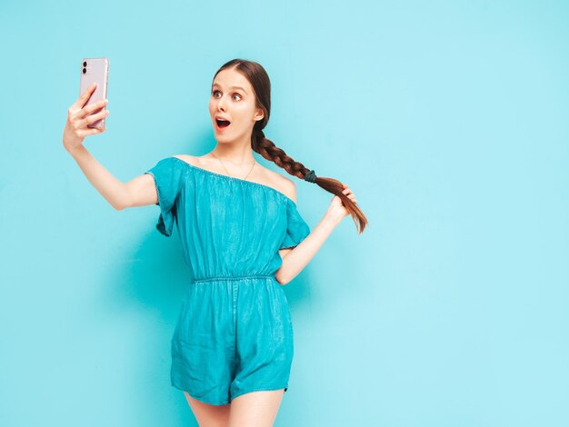 Jovem linda mulher sorridente em macacão jeans na moda de verão Mulher despreocupada sexy com penteado de cauda posando perto da parede no estúdio Modelo positivo se divertindo Alegre e feliz tomando selfie