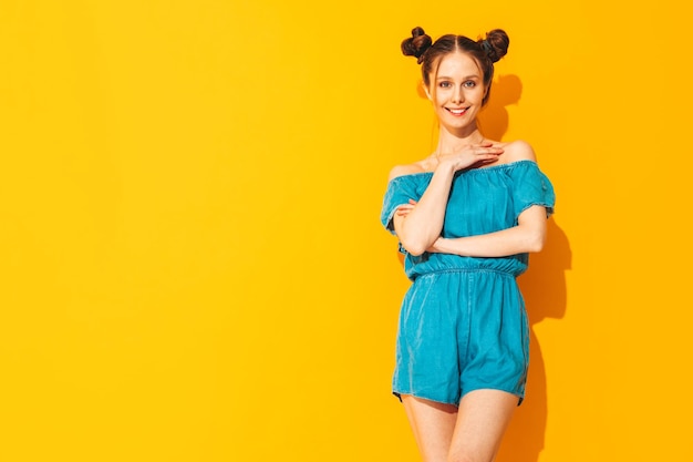 Jovem linda mulher sorridente em macacões jeans de verão na moda Mulher despreocupada sexy com penteado de dois chifres posando perto da parede amarela no estúdio Modelo positivo se divertindo isolado