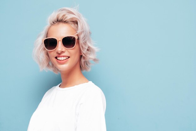 Jovem linda mulher sorridente em roupas da moda de verão Mulher despreocupada sexy posando perto da parede azul no estúdio Modelo loiro positivo se divertindo e enlouquecendo em óculos de sol