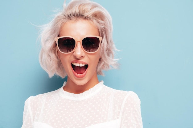 Jovem linda mulher sorridente em roupas da moda de verão Mulher despreocupada sexy posando perto da parede azul no estúdio Modelo loiro positivo se divertindo em óculos de sol Alegre e feliz gritando e gritando