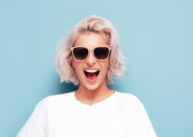 Jovem linda mulher sorridente em roupas da moda de verão Mulher despreocupada sexy posando perto da parede azul no estúdio Modelo loiro positivo se divertindo em óculos de sol gritando e gritando