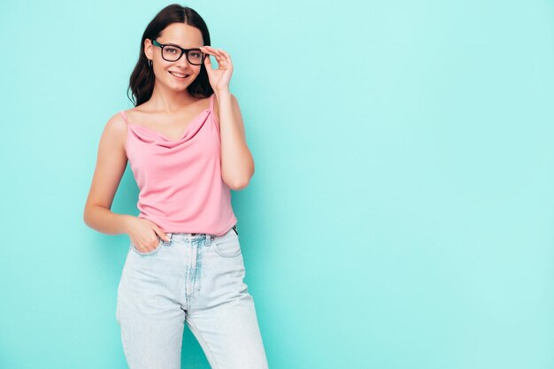 Jovem linda mulher sorridente em roupas da moda de verão Mulher despreocupada sexy posando perto da parede azul no estúdio Modelo morena positiva se divertindo e enlouquecendo Alegre e feliz em óculos