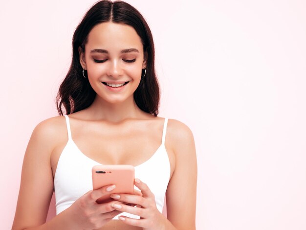 Jovem linda mulher sorridente em roupas da moda de verão Mulher despreocupada sexy posando perto da parede rosa no estúdio Modelo morena positiva olhando para a tela do celular segurando o smartphone e usando aplicativos