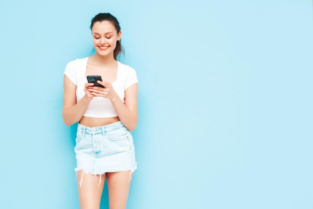 Jovem linda mulher sorridente em saia jeans na moda verão Mulher despreocupada sexy posando perto da parede azul no estúdio Modelo morena positiva olhando para a tela do celular segurando o smartphone e usando aplicativos