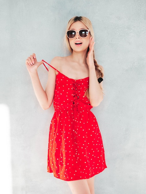 Jovem linda mulher sorridente em vestido vermelho de verão na moda Mulher loira despreocupada sexy posando perto da parede no estúdio Modelo positivo se divertindo Alegre e feliz em óculos de sol