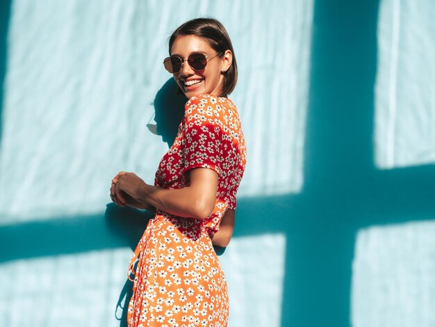 Jovem linda mulher sorridente no vestido vermelho da moda verão Mulher despreocupada sexy posando perto da parede azul no estúdio Modelo positivo se divertindo Alegre e feliz no dia ensolarado Sombra da janela