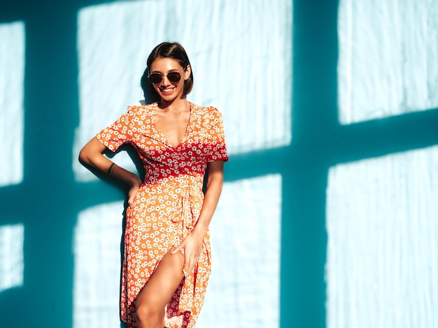 Jovem linda mulher sorridente no vestido vermelho da moda verão Mulher despreocupada sexy posando perto da parede azul no estúdio Modelo positivo se divertindo Alegre e feliz no dia ensolarado Sombra da janela