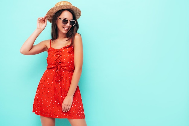 Jovem linda mulher sorridente no vestido vermelho moderno de verão mulher despreocupada sexy posando perto da parede azul no estúdio modelo morena positiva se divertindo no chapéu e óculos de sol