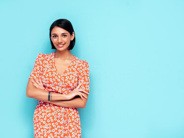 Jovem linda mulher sorridente no vestido vermelho moderno de verão Mulher despreocupada sexy posando perto da parede azul no estúdio Modelo positivo se divertindo Alegre e feliz isolado Braços cruzados