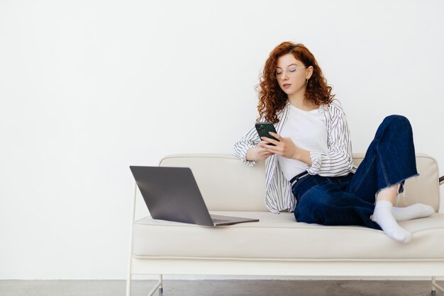 Jovem linda ruiva sentada no sofá na sala de estar e fazendo compras online no celular e laptop
