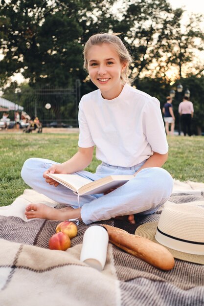 Jovem linda sorridente sentada na manta com livro olhando alegremente na câmera no piquenique no parque da cidade