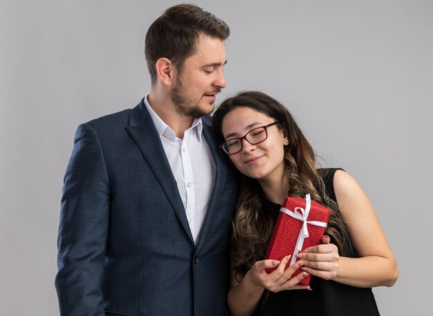 Jovem lindo casal feliz, homem e mulher com um presente, feliz e apaixonado, juntos, comemorando o dia dos namorados em pé sobre uma parede branca