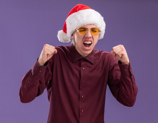 Jovem loira alegre com chapéu de Papai Noel e óculos fazendo gesto de sim com os olhos fechados, isolado na parede roxa