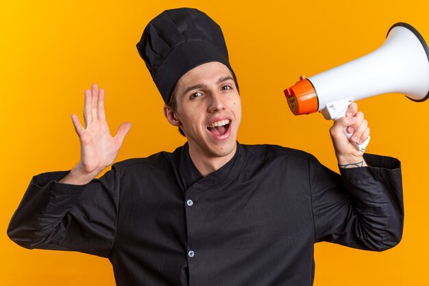 Jovem loira alegre cozinheira com uniforme de chef e boné mostrando o alto-falante e a mão vazia