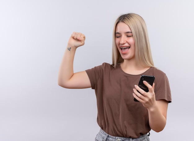 Jovem loira alegre de aparelho dentário segurando um telefone celular com o punho erguido e os olhos fechados em um espaço em branco isolado com espaço de cópia