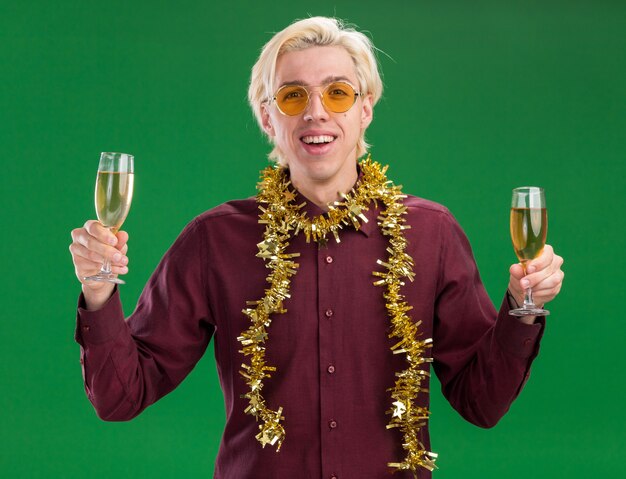 Jovem loira alegre de óculos com guirlanda de ouropel no pescoço e segurando duas taças de champanhe isoladas na parede verde