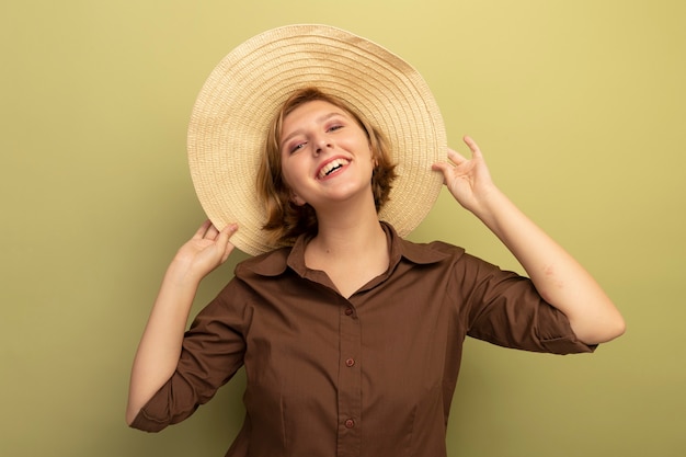 Jovem loira alegre usando um chapéu de praia, agarrando um chapéu isolado na parede verde oliva