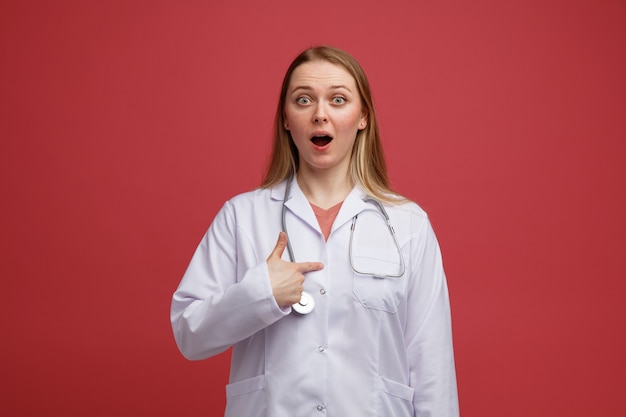 Jovem loira animada com um robe médico e um estetoscópio pendurado no pescoço e apontando para si mesma