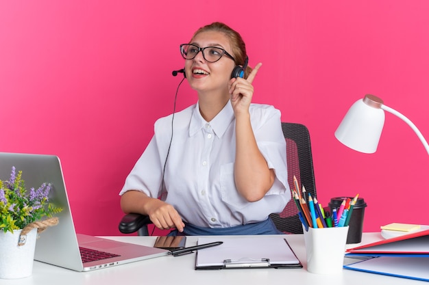 Foto grátis jovem loira animada no call center usando fone de ouvido e óculos, sentada na mesa com as ferramentas de trabalho, olhando para o lado apontando para cima, isolado na parede rosa