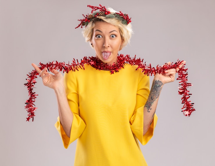 Foto grátis jovem loira brincalhona usando coroa de natal na cabeça e guirlanda de ouropel em volta do pescoço, olhando para a câmera, segurando uma guirlanda de ouropel mostrando a língua isolada no fundo branco
