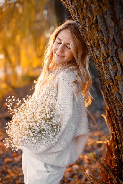 Jovem loira com flores em pé no parque outono perto do lago Mulher vestindo suéter bege e calças brancas Garota posando para uma foto em dia ensolarado