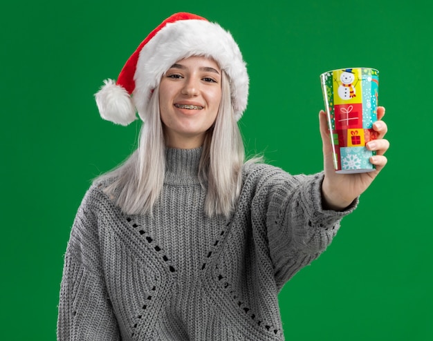 Jovem loira com suéter de inverno e chapéu de Papai Noel mostrando copo de papel clorful olhando para a câmera com um sorriso no rosto em pé sobre um fundo verde