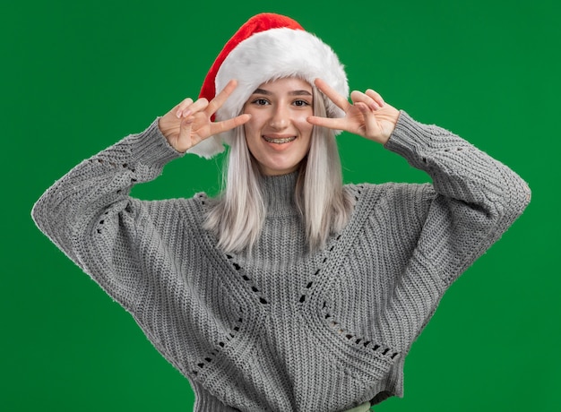 Foto grátis jovem loira com suéter de inverno e chapéu de papai noel olhando para a câmera e sorrindo alegremente mostrando o sinal v em pé sobre um fundo verde