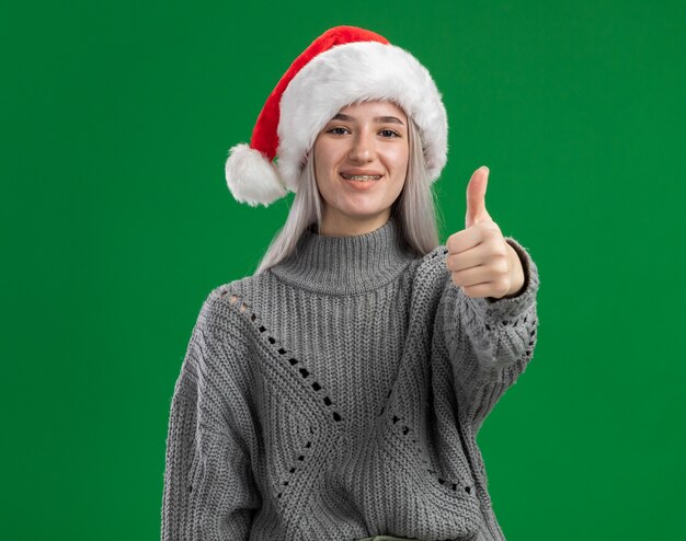 Jovem loira com suéter de inverno e chapéu de Papai Noel olhando para a câmera sorrindo e mostrando os polegares em pé sobre um fundo verde