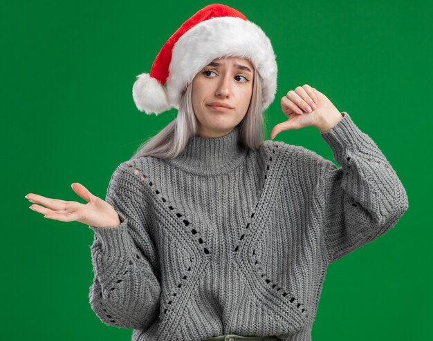 Jovem loira com suéter de inverno e chapéu de Papai Noel, parecendo confusa, mostrando o polegar para baixo, apresentando o espaço da cópia com o braço da mão em pé sobre o fundo verde