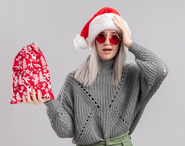 jovem loira com suéter de inverno e chapéu de Papai Noel segurando uma bolsa vermelha de Papai Noel com presentes de Natal, olhando para a câmera espantada e surpresa em pé sobre um fundo branco