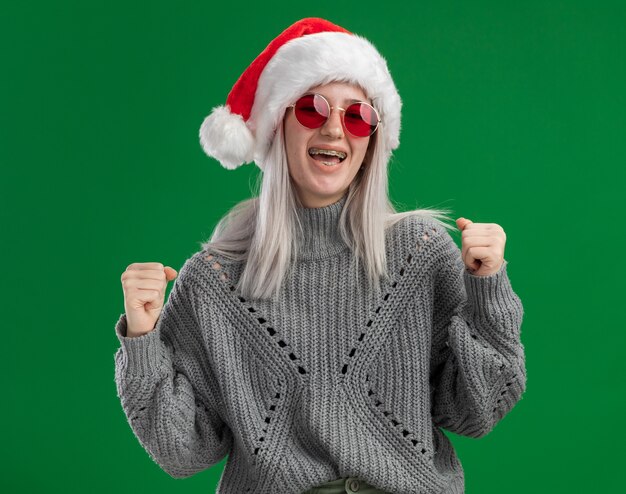 Jovem loira com suéter de inverno e chapéu de Papai Noel usando óculos vermelhos cerrando os punhos, feliz e animada em pé sobre um fundo verde