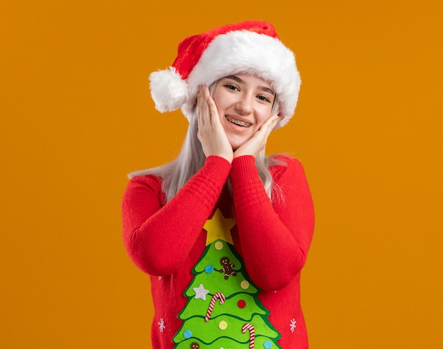Jovem loira com suéter de natal e chapéu de Papai Noel olhando para a câmera feliz e positiva sorrindo alegremente em pé sobre um fundo laranja