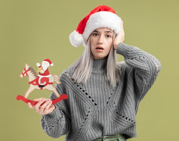 Jovem loira com um suéter de inverno e um chapéu de papai noel segurando um brinquedo de natal confuso com a mão na cabeça em pé sobre uma parede verde