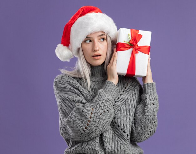 Jovem loira com um suéter de inverno e um chapéu de Papai Noel segurando um presente e parecendo intrigada em pé sobre uma parede roxa