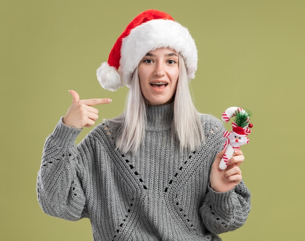 Jovem loira com um suéter de inverno e um chapéu de papai noel segurando uma bengala de doces de natal apontando com o dedo indicador para si mesma, sorrindo alegremente em pé sobre a parede verde