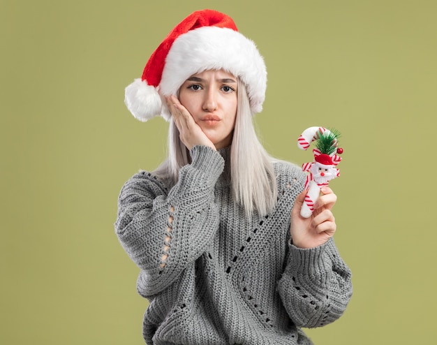 Jovem loira com um suéter de inverno e um chapéu de papai noel segurando uma bengala de doces de natal, sendo confusa em pé sobre a parede verde